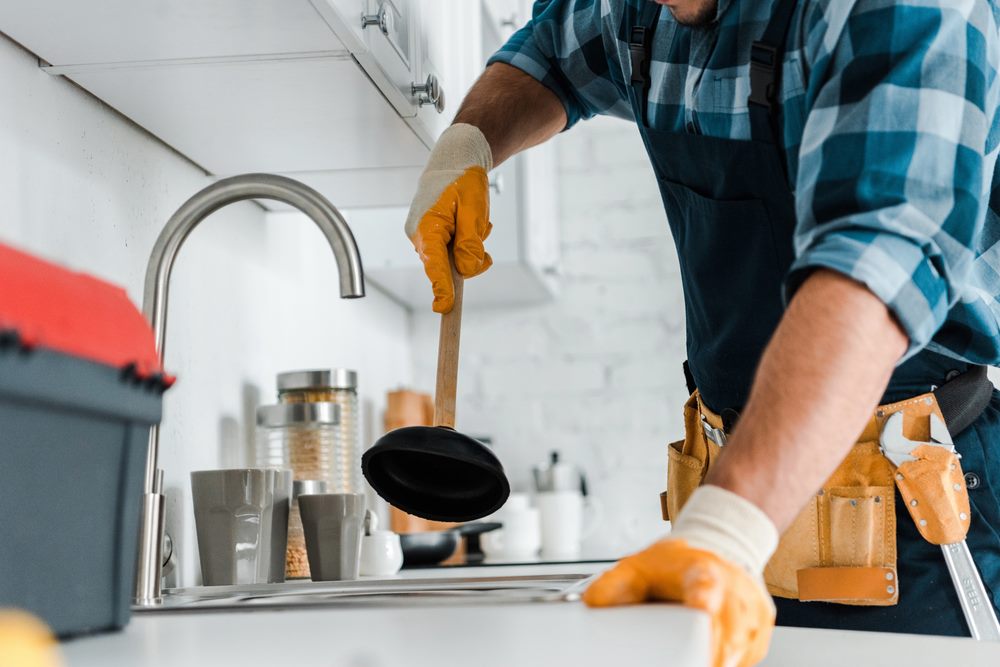 Image of a cropped view of a repairman holding a plunger in a kitchen, featuring Hero Service Pros and highlighting their motto: More Jobs. Less Hassle. Better Business.